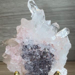 May include: A cluster of translucent white and pale pink quartz crystals featuring a prominent central dark gray mineral matrix. The crystalline formation displays sharp, pointed terminations and is supported by a gold-toned metal display stand.
