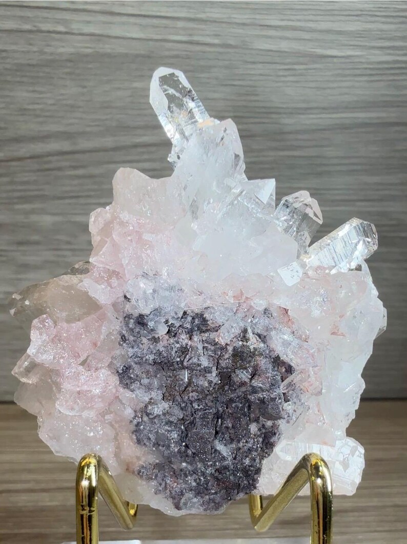 May include: A cluster of translucent white and pale pink quartz crystals featuring a prominent central dark gray mineral matrix. The crystalline formation displays sharp, pointed terminations and is supported by a gold-toned metal display stand.