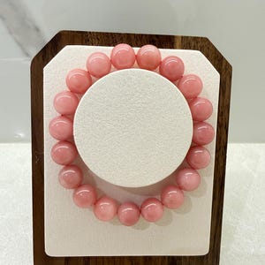 May include: A pink beaded bracelet with round beads. The bracelet is displayed on a white circular stand, set within a wooden frame. The beads are a soft, light pink color, and the bracelet is likely made of gemstones.