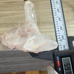 May include: A clear quartz crystal cluster with subtle pink inclusions, featuring a prominent central point. A black digital caliper is positioned against the crystal, displaying a measurement of 59.7 millimeters on its clear digital screen.