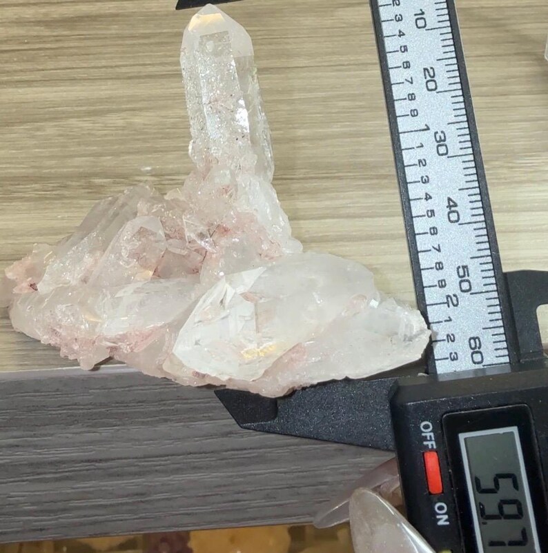 May include: A clear quartz crystal cluster with subtle pink inclusions, featuring a prominent central point. A black digital caliper is positioned against the crystal, displaying a measurement of 59.7 millimeters on its clear digital screen.