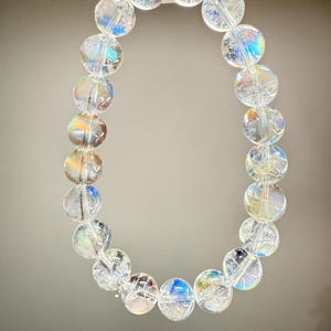May include: A clear quartz crystal bracelet with iridescent rainbow reflections. The bracelet is made of round, polished beads strung together. The beads are translucent, allowing light to pass through and create a shimmering effect. The bracelet is suspended against a neutral background.