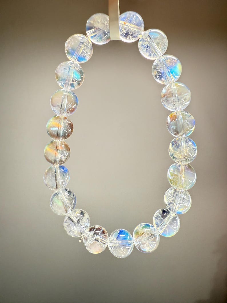 May include: A clear quartz crystal bracelet with iridescent rainbow reflections. The bracelet is made of round, polished beads strung together. The beads are translucent, allowing light to pass through and create a shimmering effect. The bracelet is suspended against a neutral background.