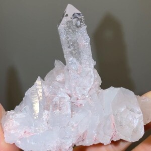 May include: A cluster of clear quartz crystals featuring a prominent central point surrounded by smaller, jagged crystalline formations. The mineral specimen displays translucent white surfaces with subtle pinkish-red inclusions embedded within the base.