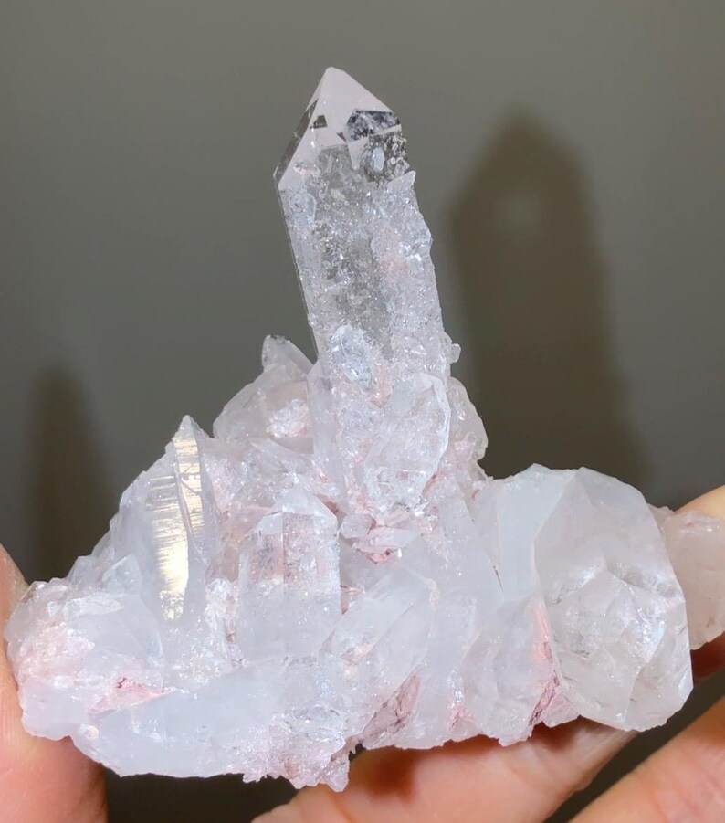 May include: A cluster of clear quartz crystals featuring a prominent central point surrounded by smaller, jagged crystalline formations. The mineral specimen displays translucent white surfaces with subtle pinkish-red inclusions embedded within the base.