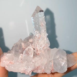 May include: A cluster of clear, translucent quartz crystals featuring a prominent central point. The mineral formation displays jagged, multifaceted surfaces with visible internal fractures and light pinkish-red mineral deposits nestled between the crystal bases.
