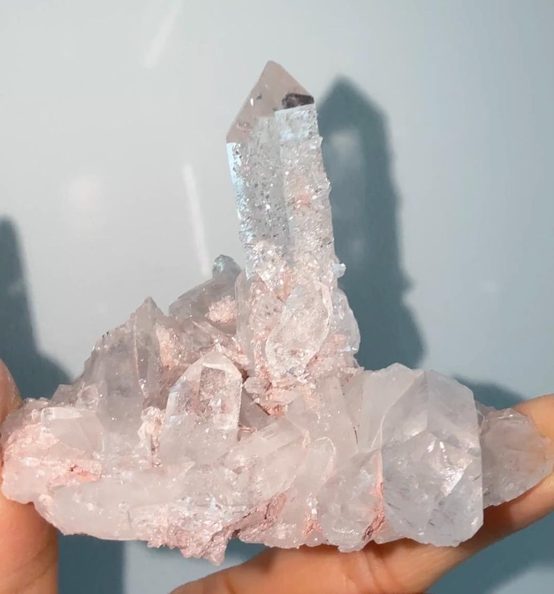 May include: A cluster of clear, translucent quartz crystals featuring a prominent central point. The mineral formation displays jagged, multifaceted surfaces with visible internal fractures and light pinkish-red mineral deposits nestled between the crystal bases.