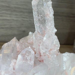 May include: A clear quartz crystal cluster featuring a prominent central point surrounded by smaller, multifaceted crystals. The mineral formation displays translucent white surfaces with distinct reddish-pink inclusions concentrated at the base.