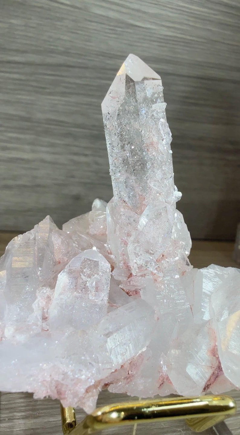 May include: A clear quartz crystal cluster featuring a prominent central point surrounded by smaller, multifaceted crystals. The mineral formation displays translucent white surfaces with distinct reddish-pink inclusions concentrated at the base.