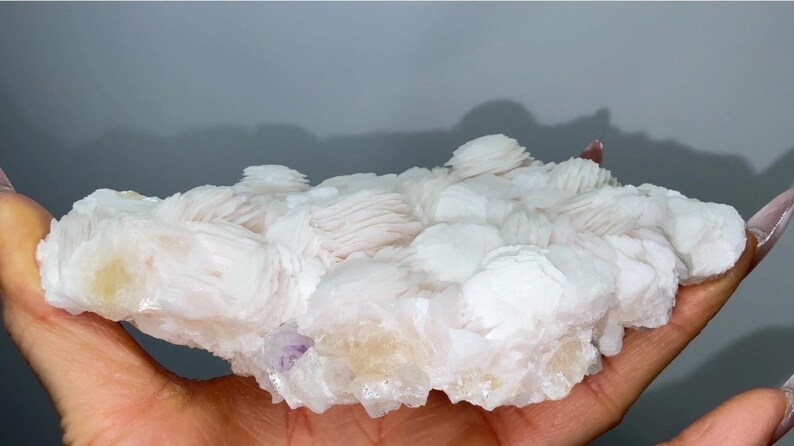 May include: A cluster of white, plate-like barite crystals forming a flat, layered formation. The mineral specimen features numerous stacked, tabular crystal blades with a pearly luster and subtle hints of pale yellow and purple coloration.