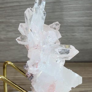 May include: A cluster of clear and translucent white quartz crystals with subtle pink inclusions, mounted on a gold-toned metal wire stand. The mineral specimen features multiple sharp, pointed terminations and a jagged, crystalline structure.