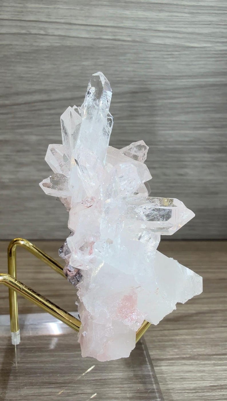 May include: A cluster of clear and translucent white quartz crystals with subtle pink inclusions, mounted on a gold-toned metal wire stand. The mineral specimen features multiple sharp, pointed terminations and a jagged, crystalline structure.