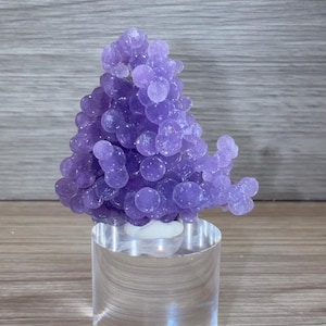 May include: A cluster of translucent purple grape agate crystals with a botryoidal formation, featuring numerous small, rounded, spherical nodules. The mineral specimen is mounted on a clear, cylindrical acrylic display stand.