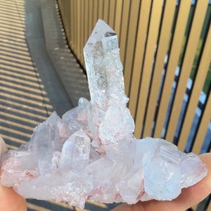 May include: A clear quartz crystal cluster featuring a prominent central point surrounded by smaller, multifaceted crystalline formations. The mineral specimen displays translucent white surfaces with subtle reddish-pink inclusions embedded within the base.
