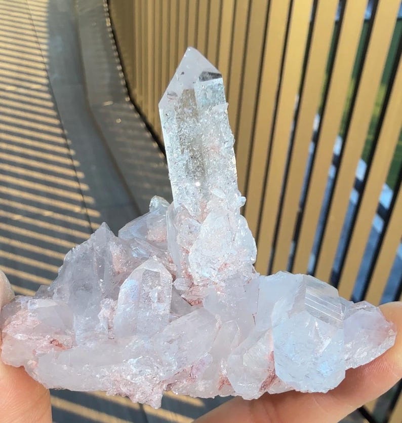 May include: A clear quartz crystal cluster featuring a prominent central point surrounded by smaller, multifaceted crystalline formations. The mineral specimen displays translucent white surfaces with subtle reddish-pink inclusions embedded within the base.