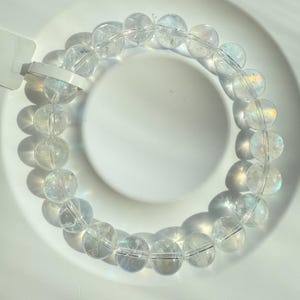 May include: A clear quartz crystal bead bracelet. The bracelet features round, translucent beads strung together. The beads have a subtle iridescent sheen, reflecting light. The bracelet is displayed on a white surface, with shadows adding depth.