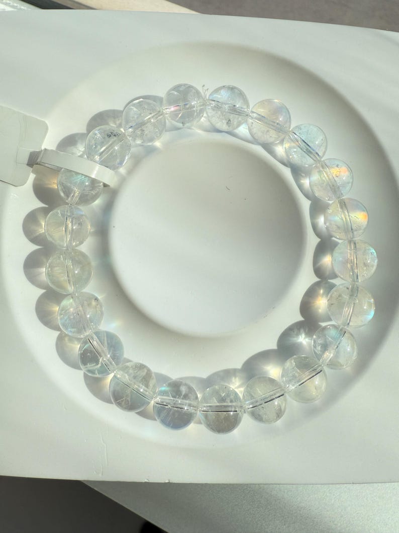 May include: A clear quartz crystal bead bracelet. The bracelet features round, translucent beads strung together. The beads have a subtle iridescent sheen, reflecting light. The bracelet is displayed on a white surface, with shadows adding depth.
