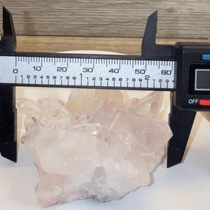 May include: A cluster of translucent, pale pink quartz crystals being measured by a black digital caliper. The display on the caliper shows a measurement of 60.2 millimeters, which is equivalent to approximately 2.37 inches in width.