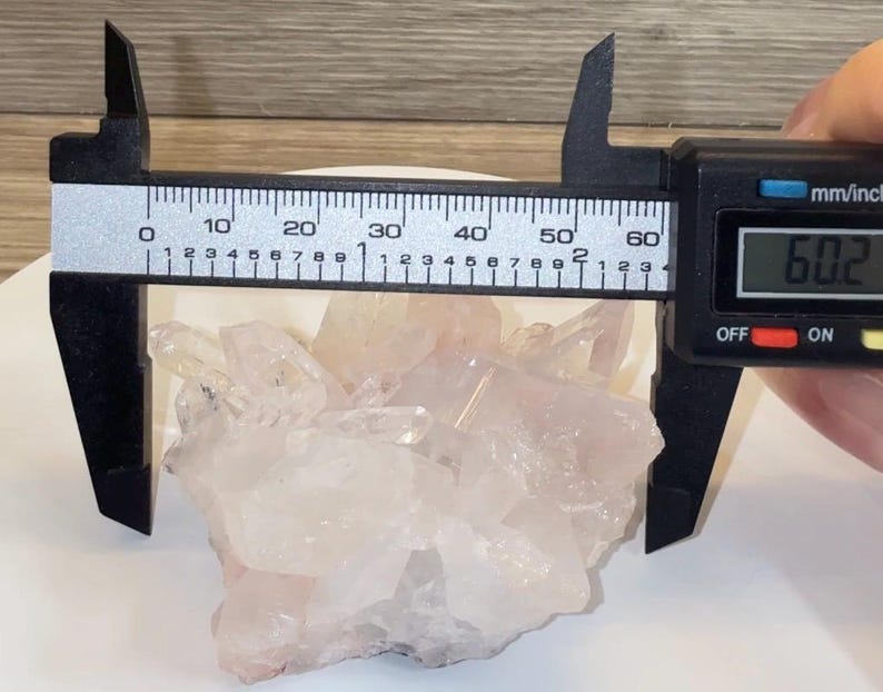 May include: A cluster of translucent, pale pink quartz crystals being measured by a black digital caliper. The display on the caliper shows a measurement of 60.2 millimeters, which is equivalent to approximately 2.37 inches in width.