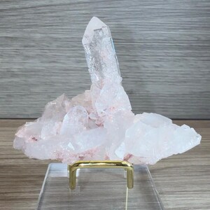 May include: A cluster of pale pink and translucent white quartz crystals featuring one prominent vertical point. The mineral specimen is mounted on a gold-toned metal wire stand that is attached to a clear, rectangular acrylic base for display.