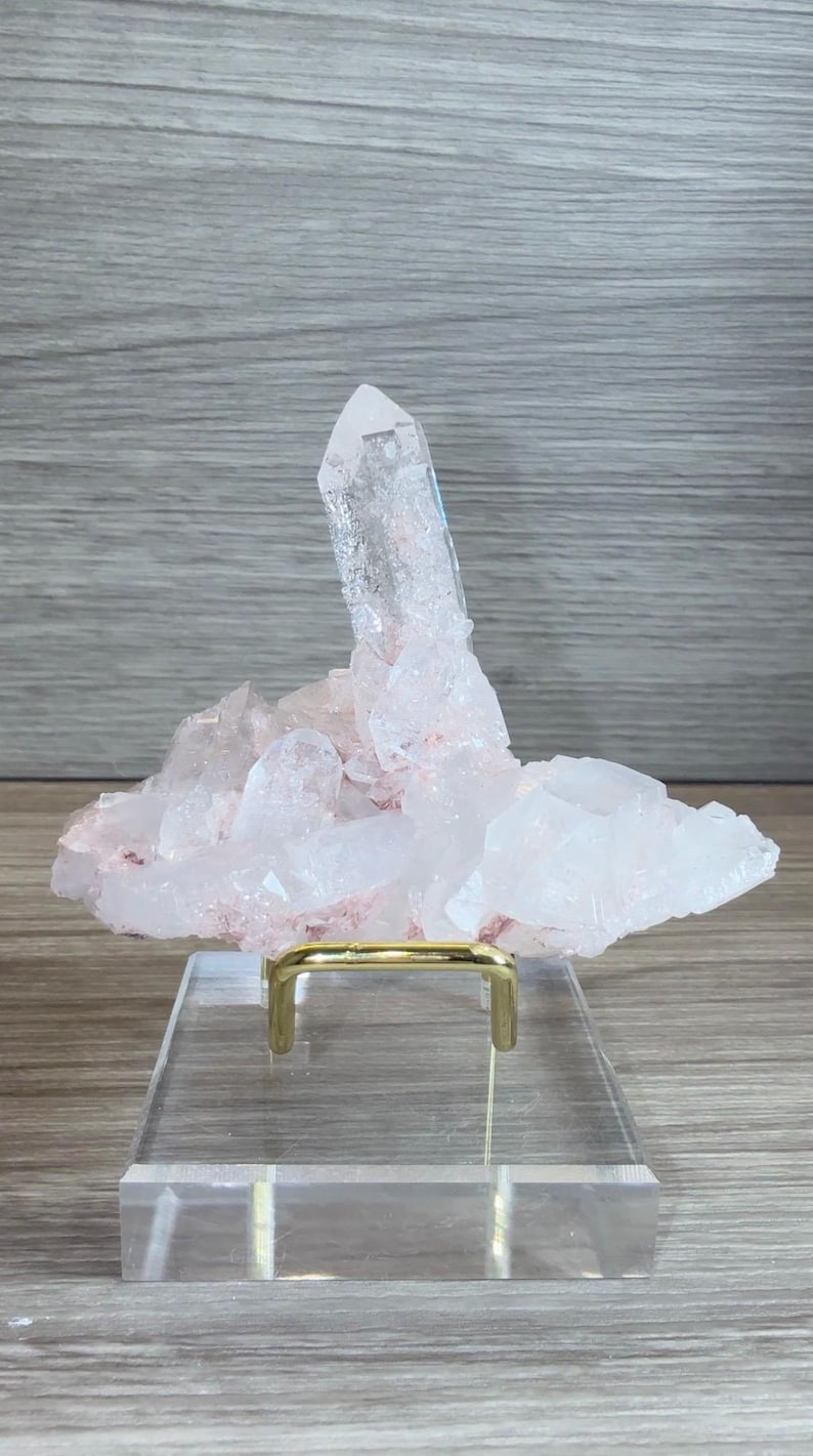 May include: A cluster of pale pink and translucent white quartz crystals featuring one prominent vertical point. The mineral specimen is mounted on a gold-toned metal wire stand that is attached to a clear, rectangular acrylic base for display.