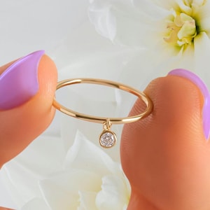 Pode incluir: Um anel fino de ouro com um pequeno pingente de pedra redonda e transparente pendurado na banda. A banda de metal é lisa e circular, e a pedra está engastada num pequeno bisel de ouro, criando uma peça de joalharia simples e delicada.