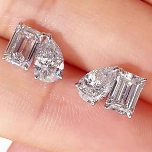 Puede incluir: Un par de pendientes de botón con una combinación de una piedra transparente de corte esmeralda y una piedra transparente en forma de pera, montadas en metal plateado. Cada pendiente muestra dos piedras facetadas lado a lado.