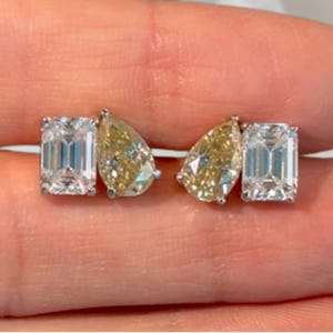 May include: A pair of earrings featuring a combination of a clear emerald-cut stone and a yellow pear-cut stone set in metal. Each earring displays a rectangular clear stone positioned next to a teardrop-shaped yellow stone, creating a symmetrical design.