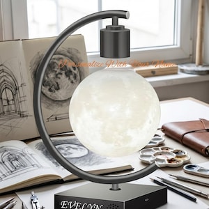 May include: A levitating moon lamp with a black base and a curved support. The moon-shaped light glows with a soft, warm light. The base is personalized with the name "EVELYN". The lamp is designed for a desk or table.