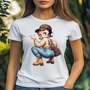 May include: White t-shirt featuring a cartoon illustration of a woman in a brown cap, tan sweater, blue jeans, and brown boots. She is pointing with her right index finger. A brown purse is on her shoulder.