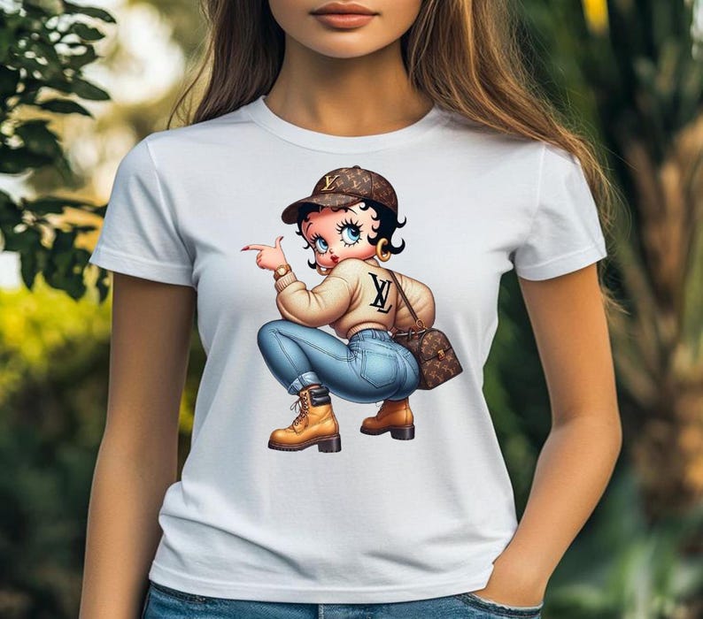 May include: White t-shirt featuring a cartoon illustration of a woman in a brown cap, tan sweater, blue jeans, and brown boots. She is pointing with her right index finger. A brown purse is on her shoulder.