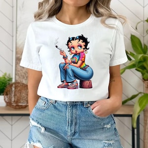 May include: White t-shirt featuring a cartoon illustration of a woman with black hair, wearing a tie-dye shirt, blue jeans, and red sandals. The character is holding a lit cigarette. The design is printed on a white t-shirt.