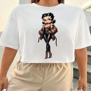 May include: White cropped t-shirt featuring a cartoon illustration of a woman in a leopard print jacket, black pants, and boots. The character has black hair and is wearing a brown jacket with black straps. The shirt is a casual, short-sleeved style.