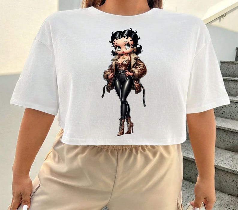 May include: White cropped t-shirt featuring a cartoon illustration of a woman in a leopard print jacket, black pants, and boots. The character has black hair and is wearing a brown jacket with black straps. The shirt is a casual, short-sleeved style.