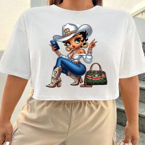 May include: White crop top featuring a cartoon cowgirl with a silver cowboy hat, blue jeans, and cowboy boots. The cowgirl is holding a phone and has a green and red handbag.