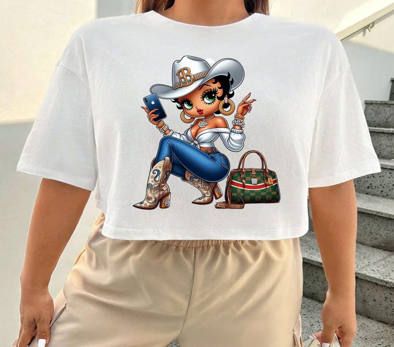 May include: White crop top featuring a cartoon cowgirl with a silver cowboy hat, blue jeans, and cowboy boots. The cowgirl is holding a phone and has a green and red handbag.