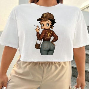 May include: White, short-sleeved crop top featuring a cartoon image of Betty Boop in a brown jacket and cap, holding a cigarette. The image has a brown purse and gray jeans. The design has a luxury brand logo.