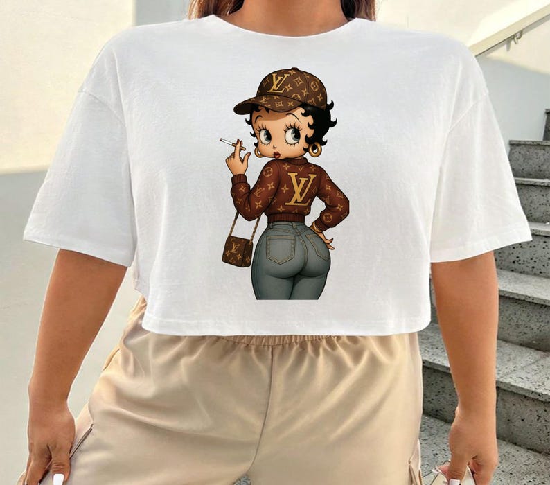 May include: White, short-sleeved crop top featuring a cartoon image of Betty Boop in a brown jacket and cap, holding a cigarette. The image has a brown purse and gray jeans. The design has a luxury brand logo.