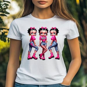 May include: White t-shirt featuring a graphic of three cartoon characters in pink tops, blue jeans, and pink boots. The middle character's top has the word "Valentine" printed on it. The characters have black hair and pink bows.