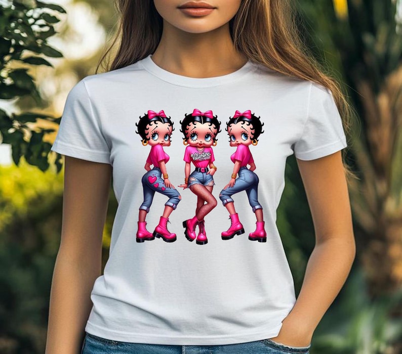 May include: White t-shirt featuring a graphic of three cartoon characters in pink tops, blue jeans, and pink boots. The middle character's top has the word "Valentine" printed on it. The characters have black hair and pink bows.
