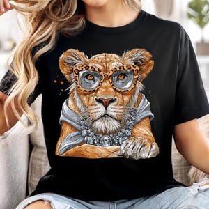 Puede incluir: Una camiseta negra con una ilustración gráfica de un cachorro de león que lleva gafas con estampado de leopardo, una bufanda gris claro alrededor del cuello y un collar grande y llamativo de tono plateado con piedras transparentes.
