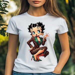 May include: White t-shirt featuring a cartoon illustration of Betty Boop in a brown and green patterned suit, red top, and green heels. The character is squatting and gesturing with one hand, wearing gold hoop earrings.