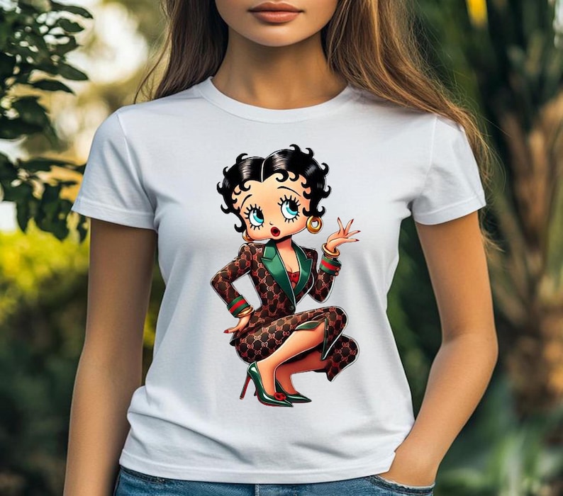 May include: White t-shirt featuring a cartoon illustration of Betty Boop in a brown and green patterned suit, red top, and green heels. The character is squatting and gesturing with one hand, wearing gold hoop earrings.