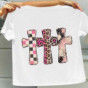 Puede incluir: Una camiseta blanca de manga corta con un estampado gráfico de tres cruces dispuestas horizontalmente. La primera cruz tiene un patrón de cuadros rosa y negro, la segunda un estampado de leopardo rosa y marrón, y la tercera es rosa con manchas negras.