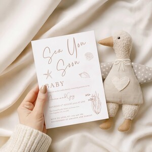 May include: A baby shower invitation with the text "Sea You Soon BABY" in a soft brown hue, and a plush duck toy. The invitation includes event details: date, time, and location. The duck toy is beige with a heart.