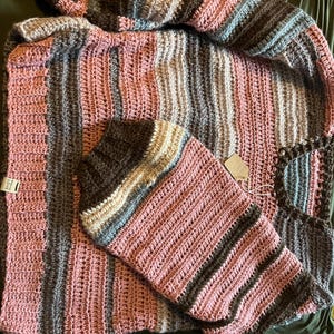 Op de afbeelding: Een handgehaakte trui met horizontale strepen in tinten oudroze, donkerbruin, lichtblauw en crème. Het kledingstuk heeft een geribbelde halslijn en boorden, met een klein wit label aan de zijkant waarop staat Made with LOVE.