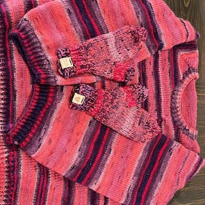 May include: A knitted sweater and matching mittens set in shades of pink, red, and purple. The sweater has a crew neck and long sleeves. The mittens have a small tag that says "LOVE". The set is made of a soft, textured yarn.
