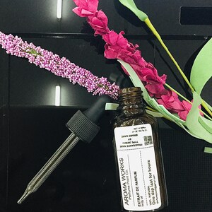 May include: Aroma Works body oil bottle with a dropper, next to pink and purple floral decorations. The label reads "EXTRAIT DE PARFUM" and "One dap last for hours". The bottle is brown glass.