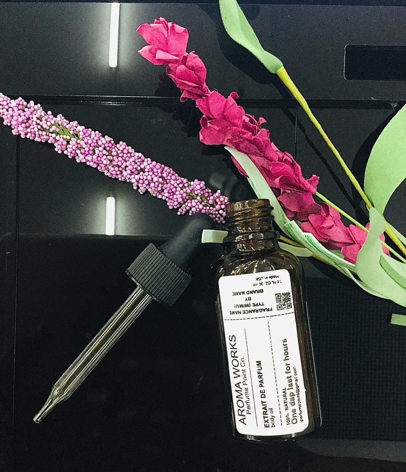May include: Aroma Works body oil bottle with a dropper, next to pink and purple floral decorations. The label reads "EXTRAIT DE PARFUM" and "One dap last for hours". The bottle is brown glass.