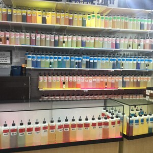 May include: A display of various colorful oils in clear bottles with black caps, arranged on shelves and in glass display cases. The bottles are labeled with numbers and contain liquids in shades of yellow, pink, blue, and more. The word "OILS" is visible.